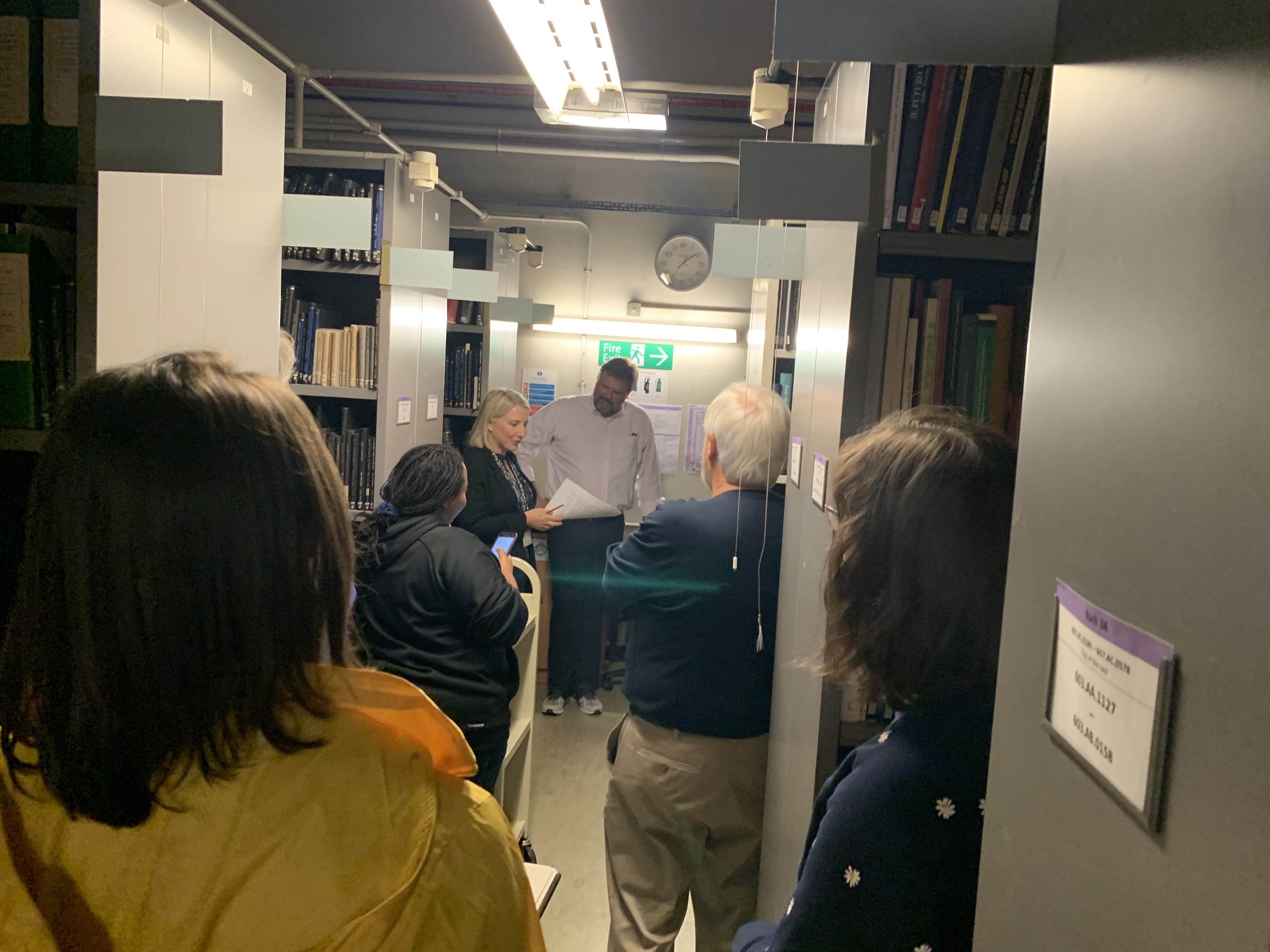 Cropped image of students and administrators listening to discussion of library and archives in tight quarters inside the stacks